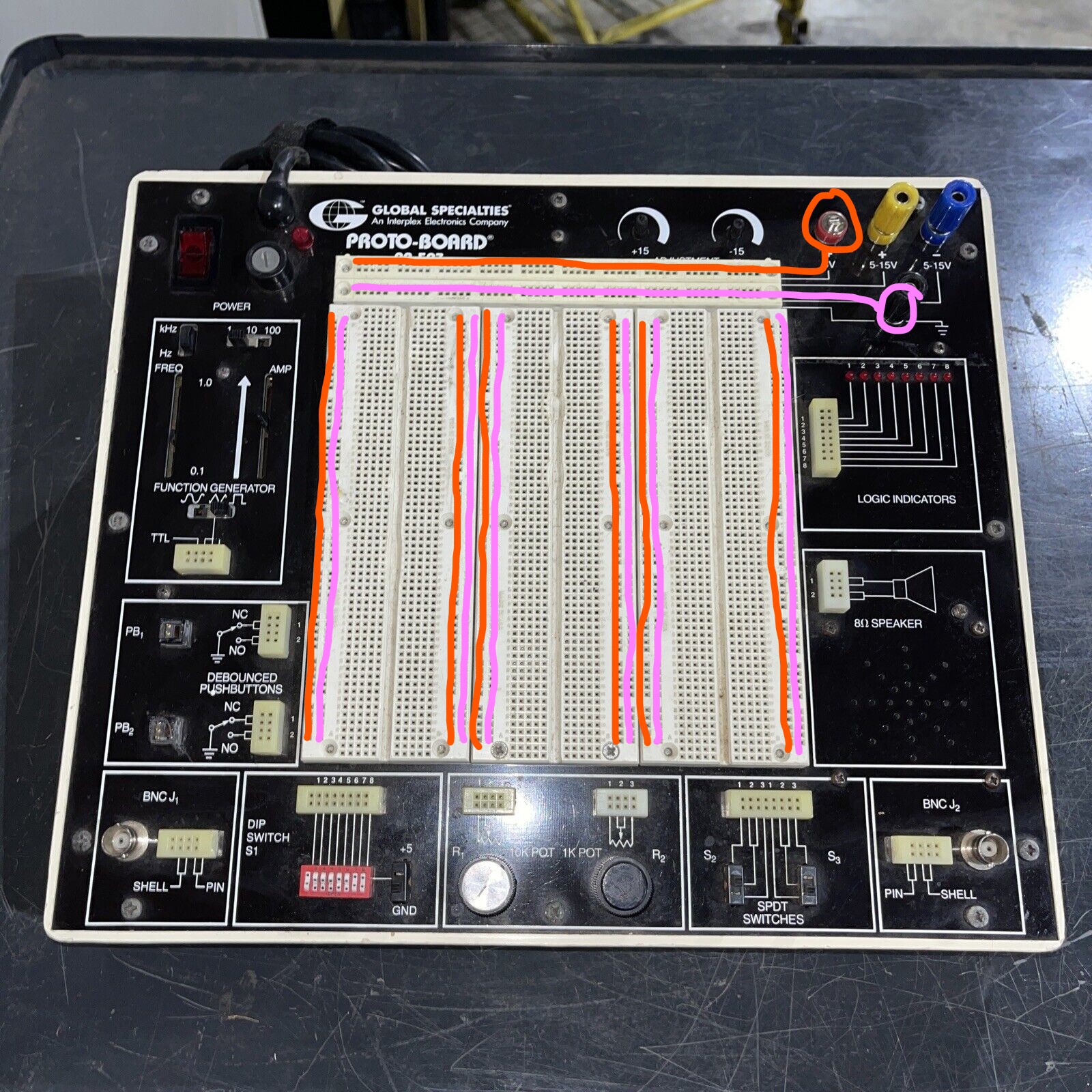 The workbench again, now showing voltage and ground connections. The +5V and GND plugs at the upper-left of the board are circled, and the horizontal socket strips for each along the top are indicated with lines (the +5V strip is the topmost two rows of sockets on the bench, and the next two rows are GND rows). From these strips, lines are drawn vertically down each of the connected columns in the protoboard section, indicating how our workbenches are pre-wired to distribute power and ground connections. Specifically, for each of the 6 pairs of vertical rails, the left-hand rail is connected to +5V and the right-hand rail is connected to ground.