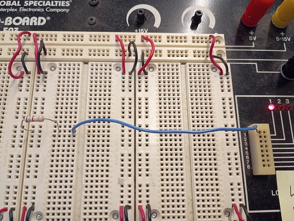 A picture of the same setup as before, except this time, a blue wire is plugged into another hole in the same row of 5 holes as the resistor, with the other end of the blue wire reaching across horizontally to the logic indicators section and plugged into one of the holes there. The logic indicator light corresponding to the place the blue wire is plugged into is lit up.