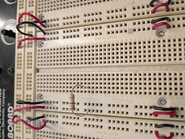 A picture of a resistor plugged into the workbench. The left leg goes into one of the square holes in a vertical column which has a red wire at the top. The right leg goes into a nearby hole in a row of 5 that’s next to one of the vertical trenches in the board.