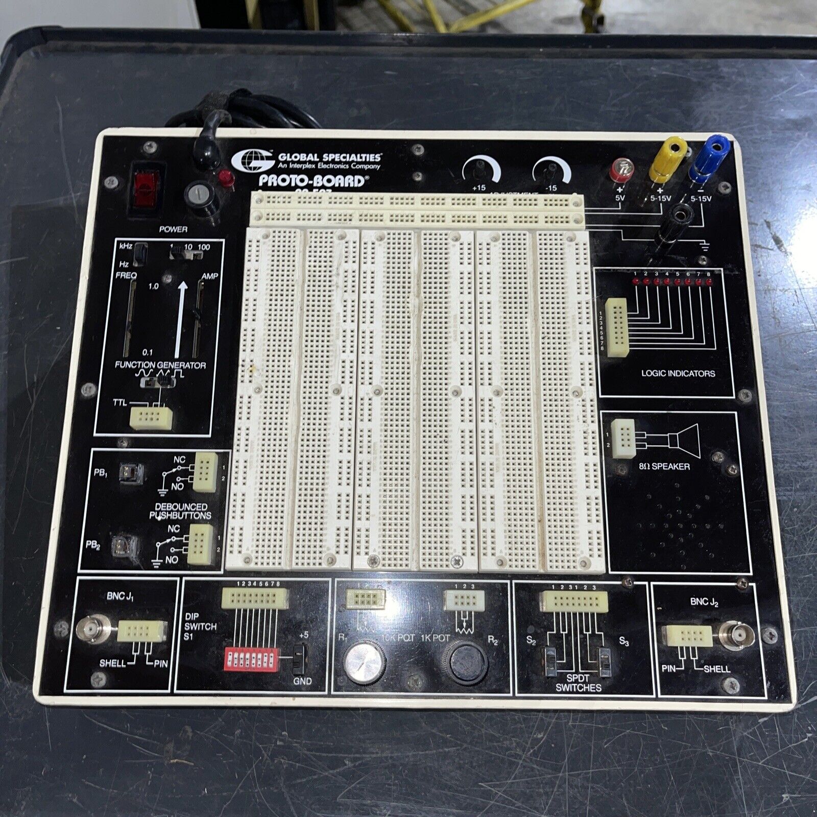One of our older-model workbenches, with voltage & ground sources at the top right, logic indicators below that, push buttons at the bottom left, logic switches below and slightly right from the push buttons, and a big area of protoboards in the middle. Voltage and ground sources are red and black colored knobs; logic indicators are 8 little LED lights with plastic sockets for connecting to them; push buttons are buttons that pop back out when you release them, along with plastic connector sockets, and logic switches are switches that can be toggles on or off, with plastic connector sockets. The protoboards are pieces of white plastic with lots of sockets in them: two columns of sockets down each side, and many rows of sockets split into two half-rows in the middle. Each socket is just big enough to plug in the tip of one of the wires we’re using. There are 6 protoboards packed edge-to-edge in a 3-wide-by-2-high setup; each is taller than it is wide so the whole setup is squarish (but slightly taller than it is wide).