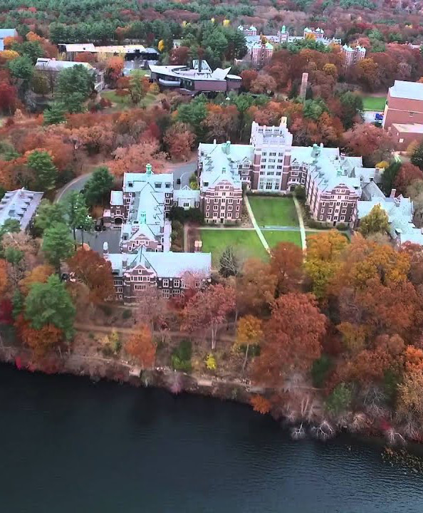 An aerial photo of the Wellesley College campus including a brick dormitory surrounding a grassy quad and plenty of trees showing red and orange Fall foliage; a lake is visible along the bottom of the image.